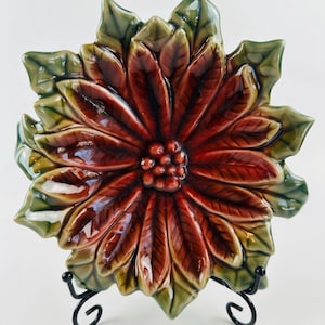 May include: A ceramic poinsettia-shaped plate with a glossy finish. The plate features deep red petals with a brown centre and green leaves. It is displayed on a black metal stand, showcasing its festive design.