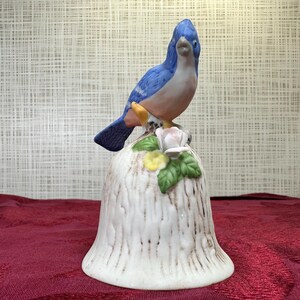 Puede incluir: Campana de cerámica con un pájaro azul posado sobre una base en forma de tocón de árbol. El pájaro tiene plumas azules y melocotón. La base es blanca con detalles marrones y adornada con una rosa rosa, flores amarillas y hojas verdes. Un artículo decorativo.