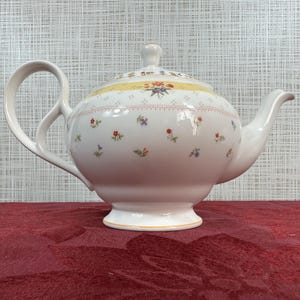 Puede incluir: Tetera de cerámica blanca con asa y boquilla curvadas. La tetera está decorada con pequeños estampados florales de colores y una banda amarilla alrededor de la tapa. La tetera está sobre una superficie roja.