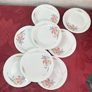 Puede incluir: Un juego de ocho platos de cerámica blanca. Cada plato presenta un diseño floral con acentos rojos, naranjas y azules. Una fina línea gris curva y una hoja dorada acentúan el diseño floral. Los platos están dispuestos sobre una superficie roja estampada.