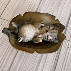 Puede incluir: Una figurita de cerámica de un gato atigrado durmiendo sobre un plato de cerámica marrón en forma de hoja. El gato está enroscado con las patas metidas debajo de la barbilla.