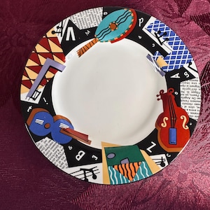 May include: A black and white ceramic plate with a colorful design featuring musical notes, a drum, and a violin. The plate has a white center and a black rim.