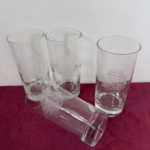 May include: Four clear glass tumblers with a ship design etched on the side. The glasses are cylindrical with a slightly flared top. The ship design is a detailed illustration of a sailing vessel.