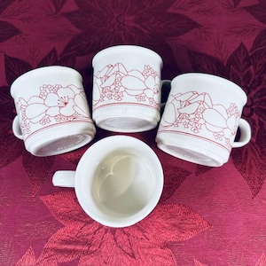 May include: Four white ceramic mugs with a red floral design. The mugs have a cylindrical shape with a handle on the side. The floral pattern is a continuous band around the top of the mugs. The mugs are arranged on a red floral patterned surface.