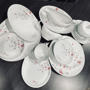 Puede incluir: Un conjunto de vajilla de porcelana blanca con un delicado estampado floral en rojo y gris. El juego incluye platos, cuencos, una salsera y una fuente cubierta, todos con ribete plateado.