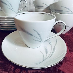 May include: White porcelain teacups and saucers with a silver rim and a wheat stalk design. The teacups have a handle and are stacked with other teacups and saucers. The set is on a red surface.