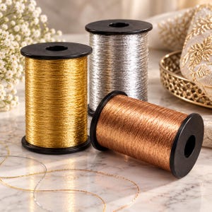 May include: Three cylindrical spools of metallic thread in gold, silver, and copper colors. Each spool features a black plastic core and circular end caps. The thread is wound tightly around the center of each spool, with loose strands extending.
