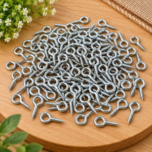 May include: A large collection of silver-coloured metal screw eyes, each featuring a circular loop at one end and a threaded screw shank at the other. These hardware fasteners are arranged in a dense pile, showing their metallic finish and shape.