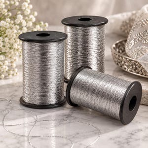 May include: Three cylindrical spools of metallic silver thread, each featuring black plastic ends. Two spools stand upright while one lies horizontally in the foreground. A thin, loose strand of the same silver thread curls across the surface.
