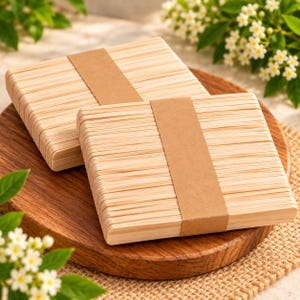 Könnte beinhalten: Zwei rechteckige Stapel aus hellen Holzstäbchen für Bastelarbeiten, die jeweils mit einem glatten, hellbraunen Papierband in der Mitte zusammengehalten werden. Die Stäbchen sind in parallelen Reihen angeordnet und zeigen eine natürliche Holzmaserung.