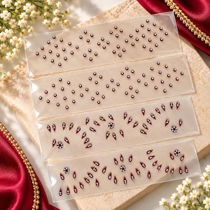 May include: Four clear adhesive sheets featuring decorative bindi stickers. The top two sheets display small round maroon gems in diamond patterns. The bottom two sheets feature maroon teardrop-shaped gems and circular floral-style accents.