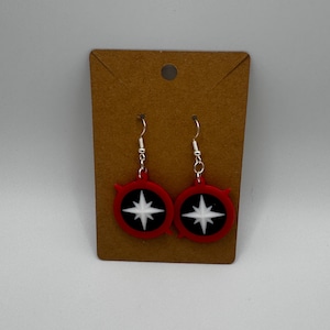 Puede incluir: Un par de pendientes de estrella roja y blanca con ganchos plateados. Los pendientes son redondos y tienen un contorno negro.