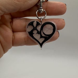 May include: A heart-shaped keychain with a black tribal design. The keychain is attached to a silver-colored clasp. The heart is clear with a black outline and design.