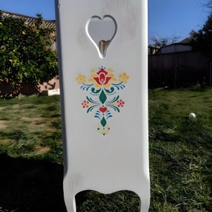 May include: A white wooden headboard with a heart-shaped cutout at the top. The headboard features a colorful floral design in red, yellow, green, and blue. The headboard is painted white and has a distressed finish.
