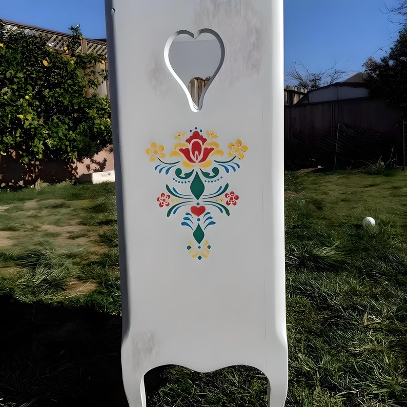May include: A white wooden headboard with a heart-shaped cutout at the top. The headboard features a colorful floral design in red, yellow, green, and blue. The headboard is painted white and has a distressed finish.