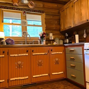 May include: A rustic kitchen with orange cabinets adorned with birch branch handles and decorative floral accents. The upper cabinets are wooden, and the room features a window with a view of trees. Decorative plates hang on the wall, adding a touch of charm.