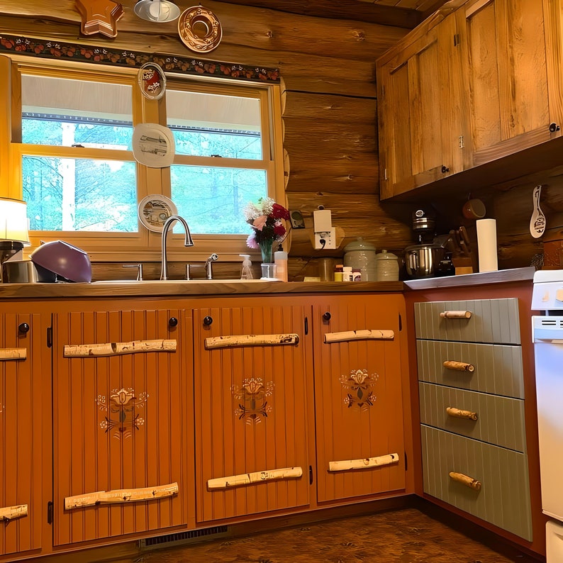 May include: A rustic kitchen with orange cabinets adorned with birch branch handles and decorative floral accents. The upper cabinets are wooden, and the room features a window with a view of trees. Decorative plates hang on the wall, adding a touch of charm.