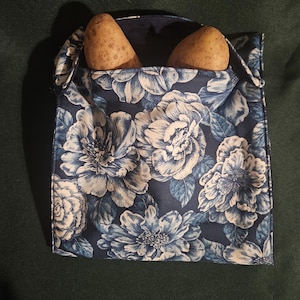 Könnte beinhalten: Eine Stofftasche mit blauem Blumenmuster, in der sich zwei Kartoffeln befinden. Das Muster der Tasche zeigt große weiße und blaue Blumen auf einem dunkelblauen Hintergrund. Die Tasche liegt auf einer dunkelgrünen Oberfläche.