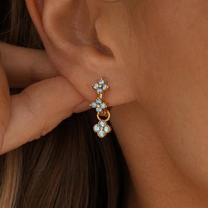 May include: A gold-toned hoop earring featuring a vertical drop design with two small, four-petaled flower charms. Each flower is set with four light blue stones arranged in a clover-like pattern, suspended from a small gold hoop.
