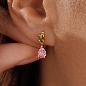 Puede incluir: Pendientes colgantes dorados con una gema verde de corte ovalado y una gema rosa en forma de pera. Los pendientes se llevan en una oreja, con una mano tocando la oreja. Rose Meanie.