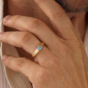 May include: A gold band ring featuring a single square-cut light blue gemstone set in the centre. The ring has a smooth, matte finish and a flat profile, showcasing a clean design that sits securely on the finger with a polished metal setting.