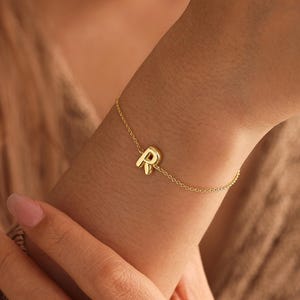 Op de afbeelding: Een goudkleurige kettingarmband met een enkele, centraal geplaatste letter R-bedel. De fijne metalen ketting wikkelt zich om de pols en toont een gepolijste afwerking op de letterhanger. Het ontwerp is eenvoudig en bestaat uit een dunne schakelketting.