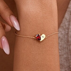Könnte beinhalten: Ein goldfarbenes Kettenarmband mit einem geteilten Herzanhänger. Eine Hälfte des Herzens ist ein tropfenförmiger roter Edelstein, die andere Hälfte ist eine glatte goldfarbene Oberfläche mit der eingravierten Großbuchstaben N, verbunden durch eine feine Kette.