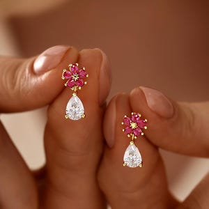 May include: A pair of gold earrings featuring a flower-shaped stud with five pink marquise-cut stones and a central accent, suspending a single clear pear-shaped drop stone. The design is delicate, showcasing vibrant pink and clear white gemstones.
