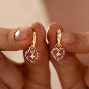 May include: A pair of gold hoop earrings featuring heart-shaped charms. Each heart is outlined with small pink stones and contains a four-pointed star cutout in the centre. A single clear stone is set into the top of each gold hoop earring.