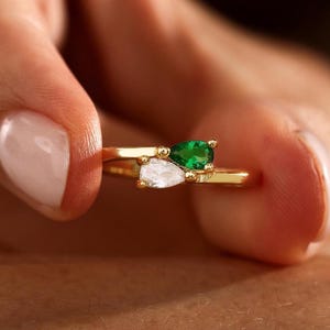 May include: A gold bypass ring featuring two pear-shaped stones set in a delicate band. One stone is a vibrant green, while the other is a clear, colourless stone. The ring has a polished metallic finish and a minimalist, open-ended design.