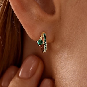May include: A gold hoop earring featuring a single prong-set round green gemstone and a row of small pavé-set green stones along the outer edge. The earring is secured in the earlobe, showcasing a polished metallic finish and vibrant green accents.