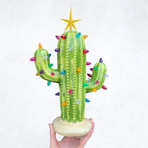 May include: A green ceramic cactus with a star on top and colorful lights strung around it. The cactus is decorated with a white base.