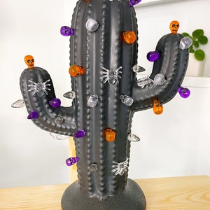 Puede incluir: Luz con forma de cactus negro con decoraciones de calaveras y arañas de color naranja, morado y blanco.
