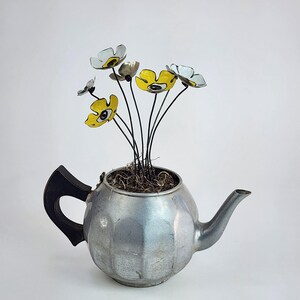 May include: A silver metal teapot used as a vase, displaying a collection of handcrafted metal flowers. The flowers feature yellow and white petals with black centers. The teapot has a black handle.