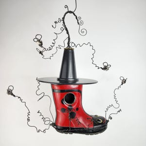 Może przedstawiać: Kapryśna, ręcznie robiona rzeźba przedstawiająca czerwoną gumową kalosz z czarnymi akcentami, czarny kapelusz i okrągłe rondo. Kalosz ma okrągły otwór i jest ozdobiony czarnymi malowanymi wzorami. Czarny drut rozciąga się od kalosza i kapelusza.