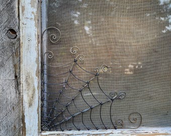 Op de afbeelding: Een zwart metalen draad spinnenweb ontwerp is bevestigd aan een wit geschilderd houten raamkozijn. Het web is gemaakt van draad en heeft een rustieke uitstraling.