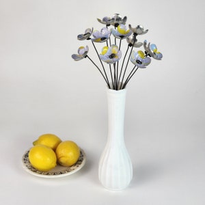 metal flowers with yellow and light blue patterns and roman numerals stand in a white vase with a plate of lemons sitting near repurposed from an old metal container.