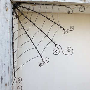 Op de afbeelding: Een zwarte metalen draadskulptuur van een spinnenweb in een hoek van een wit geschilderde muur.