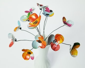 Könnte beinhalten: Ein Strauß bunter, bemalter Metallblumen in einer weißen Vase. Die Blumen sind in Blautönen, Orange, Rosa und Grün.