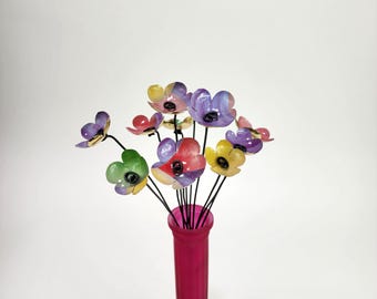 Könnte beinhalten: Ein Strauß bunter Metallblumen in einer rosa Vase. Die Blumen haben eine Vielzahl von Farben, darunter Lila, Gelb, Rot und Grün. Die Vase ist leuchtend rosa und hat eine schlanke Form. Die Stiele sind schwarz.