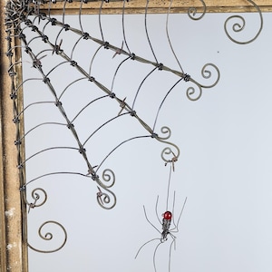 May include: A metal spider web with a red beaded spider hanging from it. The web is made of wire and is attached to a wooden frame.