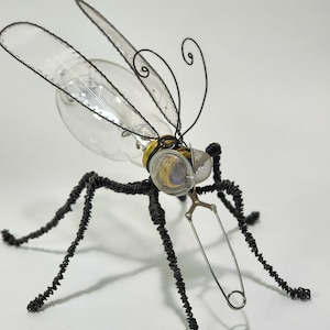 Op de afbeelding: Een zwarte draadskulptuur van een mug met een gloeilamp als lichaam en een veiligheidsspeld als proboscis.