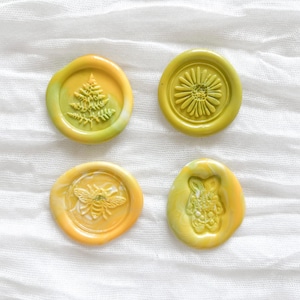 Op de afbeelding: Vier decoratieve waszegels in tinten geel en groen, elk met een uniek reliëfontwerp. De ontwerpen omvatten een varen, een madeliefje, een bij en een bloemenkonijn. De zegels zijn gerangschikt op een witte, getextureerde stof.