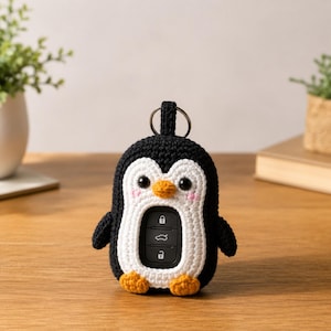 Puede incluir: Una funda de ganchillo para llave de coche con forma de pingüino en blanco y negro con una abertura central. El pingüino tiene un pequeño pico naranja, dos ojos negros, mejillas rosas bordadas y pies naranjas, con una anilla metálica arriba.