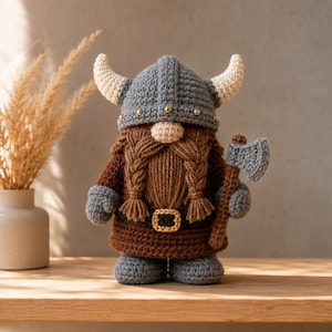 May include: A crocheted gnome figure wearing a grey horned helmet with small gold studs. The gnome has a thick brown braided beard, a brown tunic with a gold-coloured belt buckle, and grey boots. It holds a small grey crocheted battle axe.