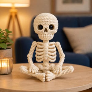 Op de afbeelding: Een crèmekleurig gehaakt skeletfiguur dat in een kleermakerszit zit. Het figuur heeft een ronde schedel met grote, gladde zwarte plastic ogen, een gestikte neusholte en een gedetailleerde ribbenkast gemaakt van garen.