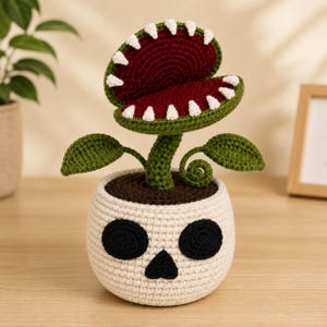 May include: A crocheted venus flytrap plant with a dark red interior, green outer leaves, and white teeth, emerging from a cream-colored crocheted pot shaped like a skull with black eye sockets and a black triangular nose cavity.