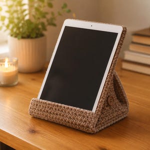 May include: A light brown, triangular crochet tablet stand featuring a textured yarn construction and a small, circular wooden button detail on the side. The stand is designed to hold a tablet device upright in a stable, angled position for viewing.