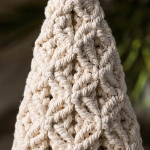 Puede incluir: Un adorno c&oacute;nico de macram&eacute; hecho de cuerda de algod&oacute;n color hueso, con un patr&oacute;n repetitivo de nudos en forma de diamante en toda la estructura. La parte superior del cono est&aacute; rematada con un peque&ntilde;o pomp&oacute;n de hilo color hueso.