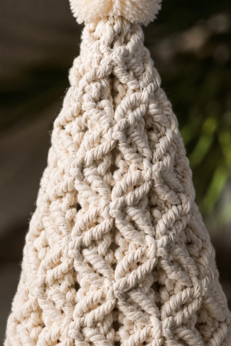 Puede incluir: Un adorno c&oacute;nico de macram&eacute; hecho de cuerda de algod&oacute;n color hueso, con un patr&oacute;n repetitivo de nudos en forma de diamante en toda la estructura. La parte superior del cono est&aacute; rematada con un peque&ntilde;o pomp&oacute;n de hilo color hueso.